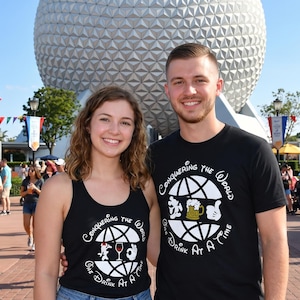 May include: A couple in matching black tops with the slogan "Conquering the World One Drink At A Time" and a globe design. The design includes a beer mug, wine glass, and Mickey Mouse hands. The background features a large silver sphere and flags.