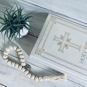 May include: A white book with gold lettering that reads "HOLY BIBLE" and a gold cross design. A white wooden bead necklace with a silver charm is lying on a white wooden surface. A small green plant is in a white pot in the upper left corner of the image.