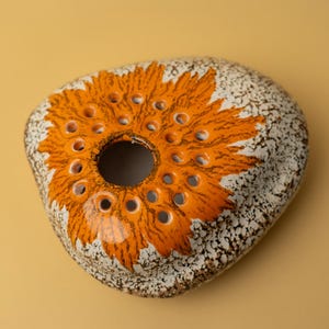 Op de afbeelding: Een keramische bloemenkikker met een oranje bloemenontwerp en een gespikkelde, gebroken witte achtergrond. De bloemenkikker heeft een centraal gat en meerdere kleinere gaten rondom.
