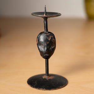 Puede incluir: Un candelero de metal negro con diseño de rostro. El candelero tiene una base redonda, un tallo central y una pequeña plataforma circular para sostener una vela. La cara es un rostro humano estilizado. El candelero mide aproximadamente 15 cm de alto.
