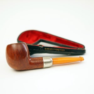 Pode incluir: Um cachimbo de madeira marrom com uma haste de âmbar amarelo e uma banda prateada. O cachimbo está em uma caixa forrada de veludo vermelho com as palavras "Guaranteed Pure Briar" impressas na caixa.
