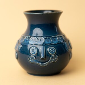 Puede incluir: Un jarrón de cerámica azul oscuro con un diseño de cara sonriente. El jarrón tiene un acabado brillante y presenta detalles en relieve alrededor de la cara y la parte superior del cuerpo. El jarrón mide aproximadamente 20 cm de altura.