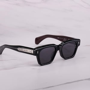 May include: Black sunglasses with a thick, rectangular frame and dark tinted lenses. The temples feature a silver-toned metallic inlay with a cross emblem. The inner side of the temple is a deep burgundy color, providing a contrast to the black exterior.
