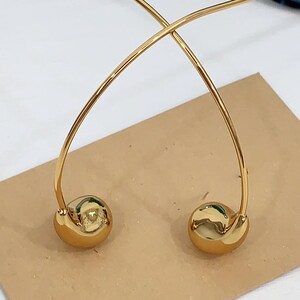 May include: A pair of gold-colored earrings. Each earring features a curved, thin, gold wire that arches over and connects to a small, round, gold-colored ball. The earrings are displayed on a brown surface.