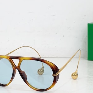 May include: Aviator-style sunglasses with a tortoiseshell frame and light blue lenses. The gold-colored arms have a unique design with a small, round accent near the ear. A green box is in the background.