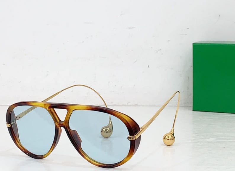 May include: Aviator-style sunglasses with a tortoiseshell frame and light blue lenses. The gold-colored arms have a unique design with a small, round accent near the ear. A green box is in the background.
