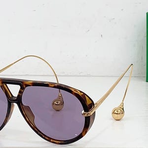 May include: Aviator-style sunglasses with a tortoiseshell frame and purple lenses. The gold-colored arms feature a unique design with a small, round accent. A green box is in the background.