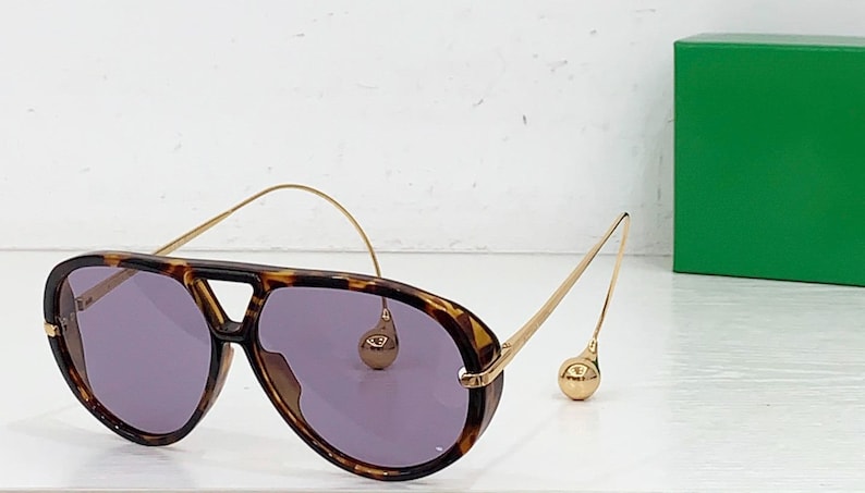 May include: Aviator-style sunglasses with a tortoiseshell frame and purple lenses. The gold-colored arms feature a unique design with a small, round accent. A green box is in the background.