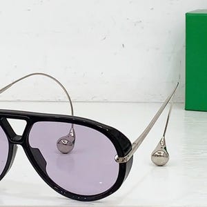 May include: Black aviator sunglasses with purple lenses and silver-tone metal accents. The sunglasses feature a unique design with teardrop-shaped embellishments and a curved top bar. A green box is in the background.