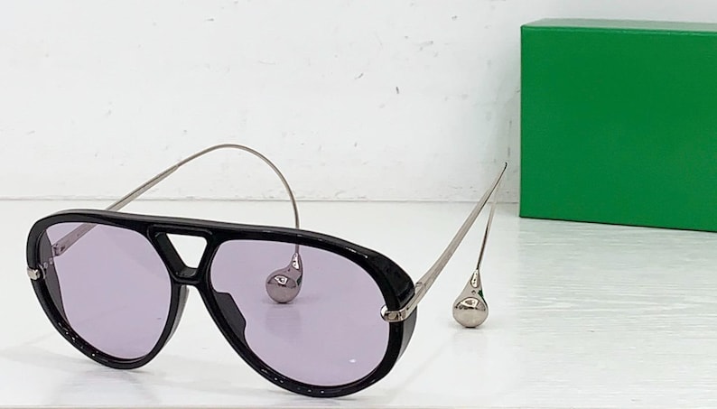 May include: Black aviator sunglasses with purple lenses and silver-tone metal accents. The sunglasses feature a unique design with teardrop-shaped embellishments and a curved top bar. A green box is in the background.
