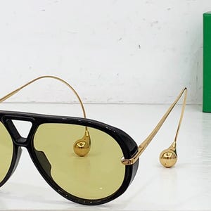 May include: Black aviator sunglasses with yellow lenses and gold-colored accents. The sunglasses have a thick black frame and a bridge. The temples are thin and gold, with a decorative gold sphere at the end.
