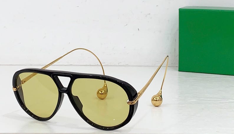 May include: Black aviator sunglasses with yellow lenses and gold-colored accents. The sunglasses have a thick black frame and a bridge. The temples are thin and gold, with a decorative gold sphere at the end.