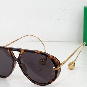 May include: Aviator-style sunglasses with a tortoiseshell frame and dark lenses. The gold-colored temples have a unique design with a small, round accent at the end. The sunglasses are set against a white background.