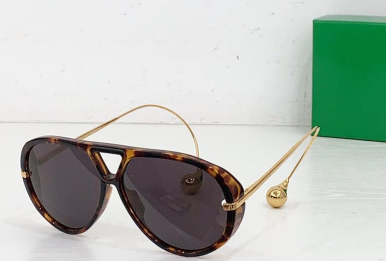 May include: Aviator-style sunglasses with a tortoiseshell frame and dark lenses. The gold-colored temples have a unique design with a small, round accent at the end. The sunglasses are set against a white background.