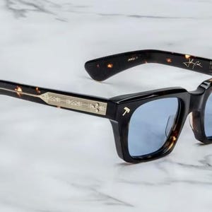 May include: Square-framed sunglasses with a black and tortoiseshell pattern. The lenses are a light blue color. The temples have a gold-colored accent. The glasses are on a white marble surface.