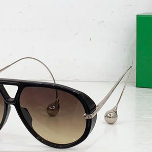 May include: Black aviator sunglasses with gradient brown lenses and silver-toned metal arms. The sunglasses have a thick black frame and a distinctive bridge design. A green box is in the background.