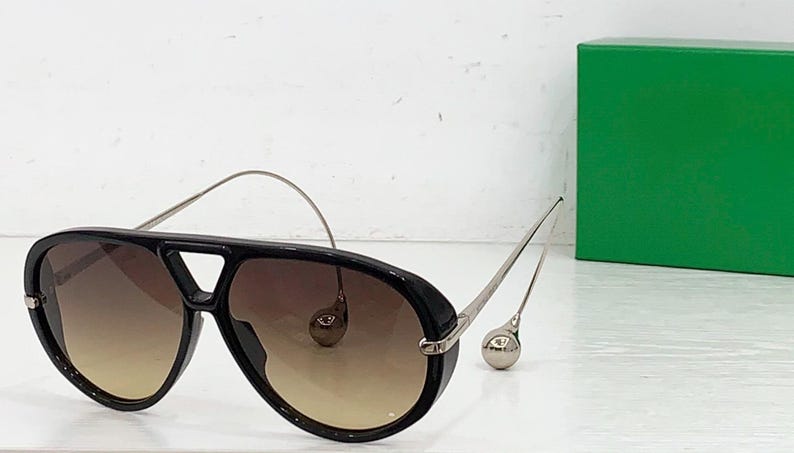 May include: Black aviator sunglasses with gradient brown lenses and silver-toned metal arms. The sunglasses have a thick black frame and a distinctive bridge design. A green box is in the background.