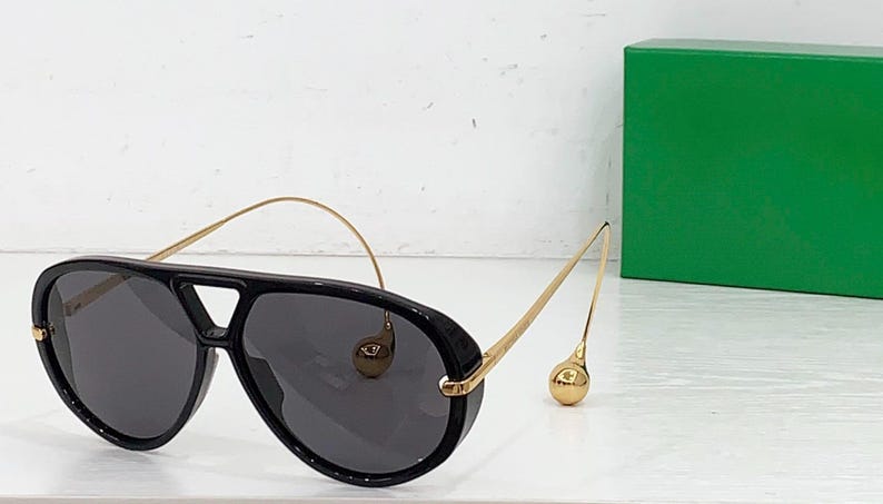 May include: Black aviator sunglasses with gold-tone metal arms and a distinctive bridge. The sunglasses have dark lenses and a rounded shape. A green box is in the background.