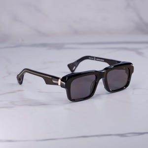 May include: Black rectangular sunglasses featuring thick frames, dark tinted lenses, and a gold-tone metal accent on the temple hinges. The temples have a distinct angular shape with a patterned cutout detail at the tips and white text printed inside.