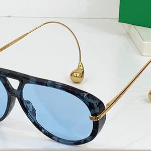 May include: Aviator-style sunglasses with a blue and black patterned frame and light blue lenses. The gold-colored temples curve down to a teardrop-shaped accent. A stylish accessory for eye protection.