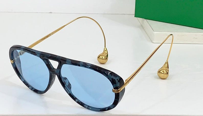 May include: Aviator-style sunglasses with a blue and black patterned frame and light blue lenses. The gold-colored temples curve down to a teardrop-shaped accent. A stylish accessory for eye protection.