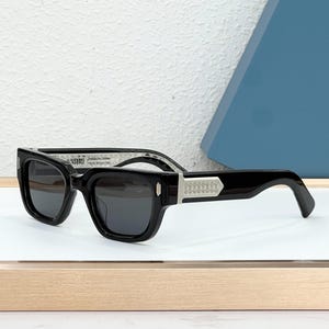 May include: Black sunglasses with a rectangular frame. The sunglasses have dark lenses and a silver decorative element on the temples. The text "AMIRI" is visible on the frame. The sunglasses are displayed on a white surface.