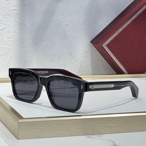 May include: Black square-framed sunglasses with dark lenses. The sunglasses have a thick frame and a silver accent on the temples. The text "JACQUESMARIE MAGE LOS ANGELES" is printed on the frame.