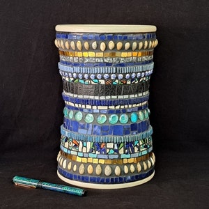 Könnte beinhalten: Eine zylindrische Keramikvase mit Mosaikdesign. Die Vase ist mit blauen, türkisfarbenen, goldenen und weißen Fliesen verziert. Die Vase steht auf einer schwarzen Oberfläche.