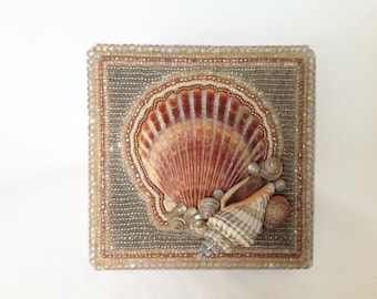 Könnte beinhalten: Eine quadratische Schachtel mit einem Perlenrahmen und einem Muschelmotiv. Die Schachtel ist mit einer großen Jakobsmuschel und mehreren kleineren Muscheln verziert.