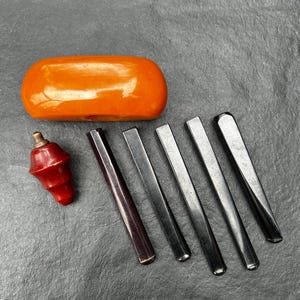 Puede incluir: Una colección de accesorios para fumar que incluye un cuenco de pipa grande, liso, ovalado y de color naranja, un componente de pipa rojo pequeño y cónico con una punta de metal color latón, y cinco boquillas de pipa negras y delgadas de varios tamaños.
