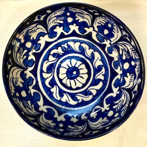 Può includere: Una ciotola rotonda in ceramica con una glassa blu scuro e intricati motivi floreali bianchi. Il design presenta un motivo floreale centrale circondato da foglie vorticose e bordi decorativi. Il bordo della ciotola è anch'esso blu scuro.