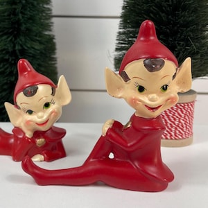 Puede incluir: Dos figuras de duendes navideños de cerámica. Los duendes están en una pose reclinada, vistiendo trajes y sombreros rojos. Tienen caras sonrientes con ojos verdes y orejas puntiagudas. El fondo incluye pequeños árboles de Navidad verdes.