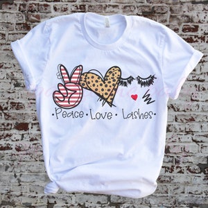 May include: White t-shirt with a peace sign, heart, and eyelashes graphic. The peace sign is red and white stripes, the heart is leopard print, and the eyelashes are black. The text "Peace Love Lashes" is printed below the graphic.
