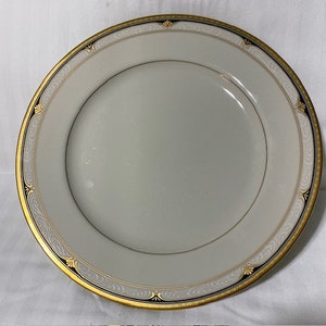 May include: A white ceramic dinner plate with a gold and black rim. The rim has a decorative pattern of scrolls and leaves.