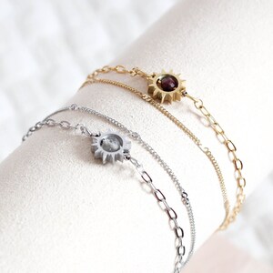 Peut inclure: Trois bracelets délicats avec des breloques en forme de soleil. L'un est doré avec une pierre centrale rouge foncé, un autre est argenté avec une pierre centrale transparente, et le troisième est doré. Les bracelets sont exposés sur une surface cylindrique blanche.
