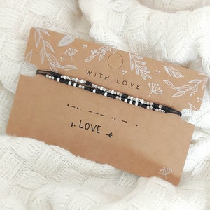 Peut inclure: Un bracelet de perles noires et blanches avec un motif en code Morse, présenté sur une carte marron avec le mot "LOVE". La carte est accompagnée d'une seconde carte avec l'inscription "WITH LOVE" et des illustrations florales. Le fond est un tissu blanc texturé.