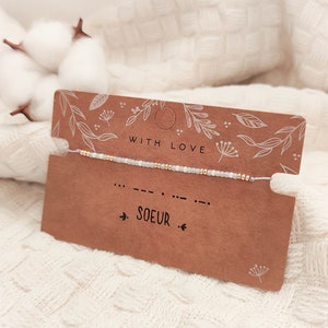Peut inclure: Un bracelet de perles délicat avec des perles blanches et dorées est présenté sur une carte marron. La carte indique "WITH LOVE" et "SOEUR". Le bracelet est sur un fond blanc avec des fleurs de coton.