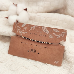 Peut inclure: Un bracelet en fil noir avec des perles dorées et noires, présenté sur une carte marron avec le code Morse pour "PAPA". La carte est ornée de motifs floraux et des mots "WITH LOVE".