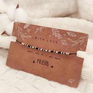 Peut inclure: Un bracelet en fil noir avec des perles dorées et noires épelant "AMI" en code Morse est présenté sur une carte marron. La carte porte les mots "AVEC AMOUR" et des motifs floraux. Le fond est un tissu blanc texturé.