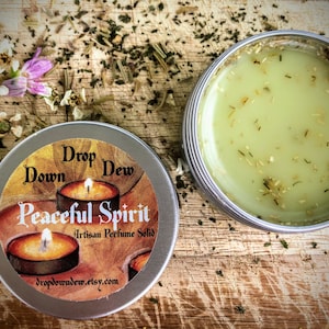 May include: A silver tin with a label that reads "Drop Down Dew Peaceful Spirit Artisan Perfume Solid" and a website address. The tin is open and shows a light green solid perfume with small green flecks.