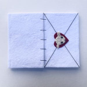 May include: A white felt notebook with black stitching along the spine. A red heart with a white bunny face is attached to the front cover. Black thread creates an envelope design on the front.