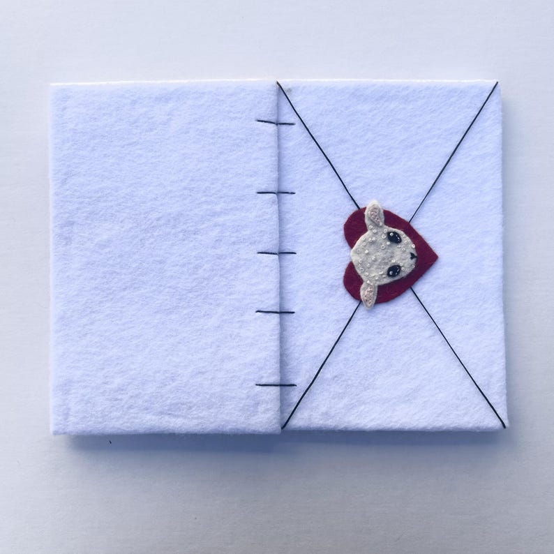 May include: A white felt notebook with black stitching along the spine. A red heart with a white bunny face is attached to the front cover. Black thread creates an envelope design on the front.