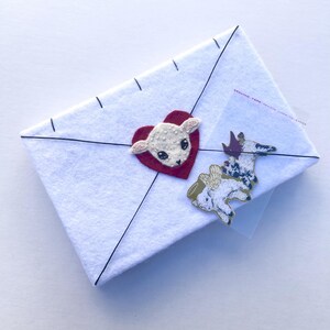 May include: A white felt envelope with black stitching and a red heart with a lamb applique. A sticker of a lamb with wings is attached to the envelope. The envelope is sealed with clear tape.