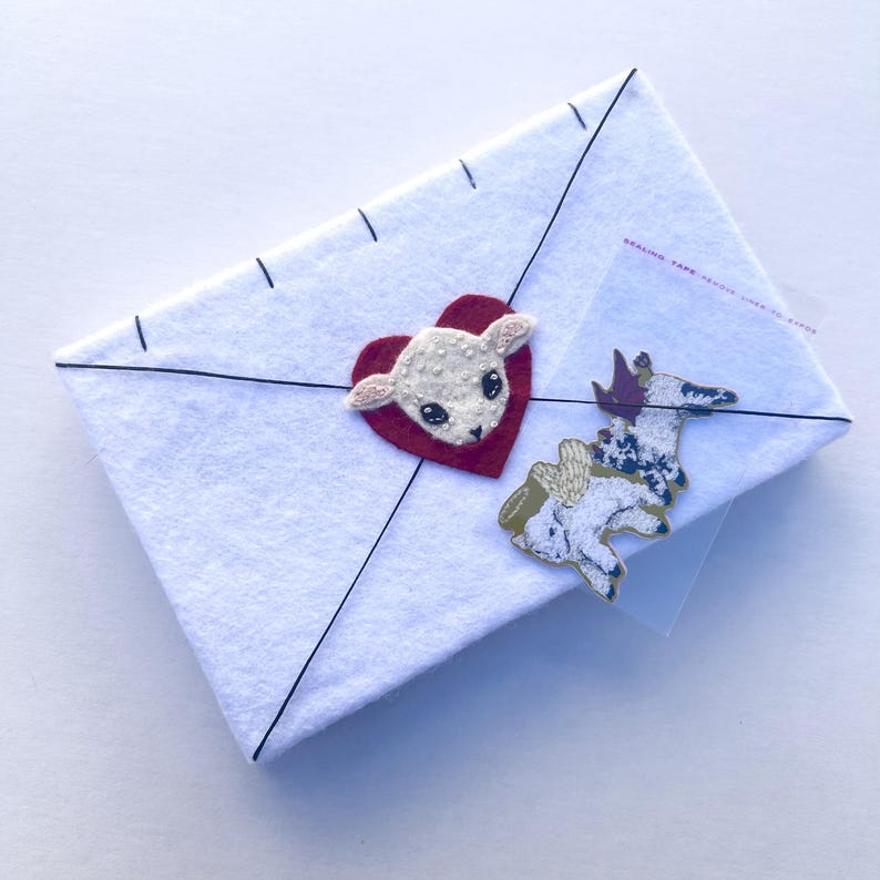 May include: A white felt envelope with black stitching and a red heart with a lamb applique. A sticker of a lamb with wings is attached to the envelope. The envelope is sealed with clear tape.