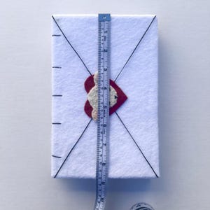 May include: A white felt envelope with a red heart and a white beaded design. The envelope is approximately 10 inches tall. Black lines create an envelope design. A blue and yellow measuring tape is placed on the envelope.