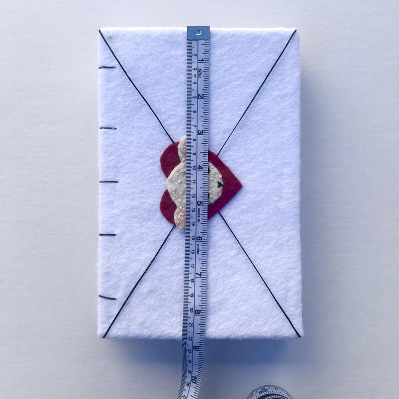 May include: A white felt envelope with a red heart and a white beaded design. The envelope is approximately 10 inches tall. Black lines create an envelope design. A blue and yellow measuring tape is placed on the envelope.