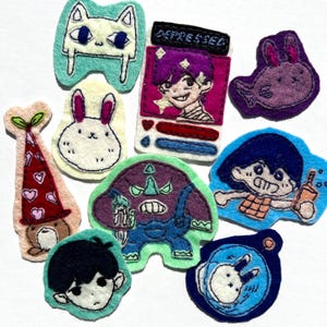 May include: A collection of eight colorful felt patches featuring various embroidered character designs, including a purple monster, a white rabbit, a character labeled DEPRESSED, and other stylized figures with distinct outlines and vibrant thread colors.