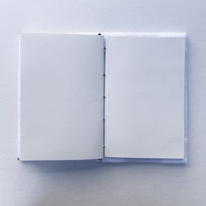 May include: An open, blank notebook with white pages. The notebook is bound with black thread and has a light blue cover. The pages are clean and ready for writing or sketching. The notebook is on a white surface.