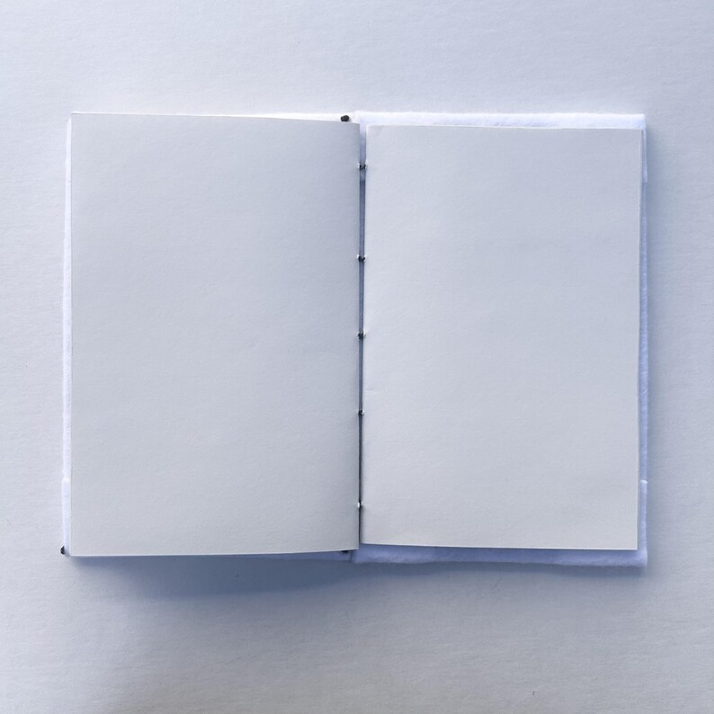 May include: An open, blank notebook with white pages. The notebook is bound with black thread and has a light blue cover. The pages are clean and ready for writing or sketching. The notebook is on a white surface.