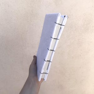 May include: A handmade journal with a light purple felt cover and white pages. The pages are bound with black thread, creating a visible stitch pattern along the spine. The journal is held upright, showcasing its rectangular shape and textured cover.
